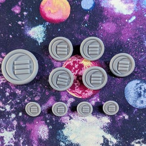 Op de afbeelding: Een verzameling grijze, ronde knopen met een maan en drie horizontale lijnen. De knopen variëren in grootte en zijn gerangschikt op een kleurrijke stof met ruimtethema. De knopen zijn gemaakt van een solide, matte materiaal.