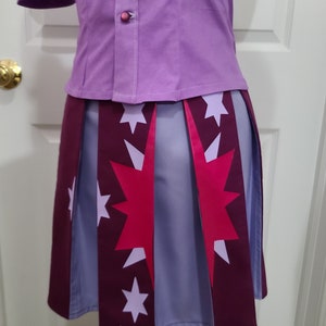 May include: A purple button-up shirt with a pink button and a pleated skirt with a pink and white star pattern. The skirt has a burgundy trim.