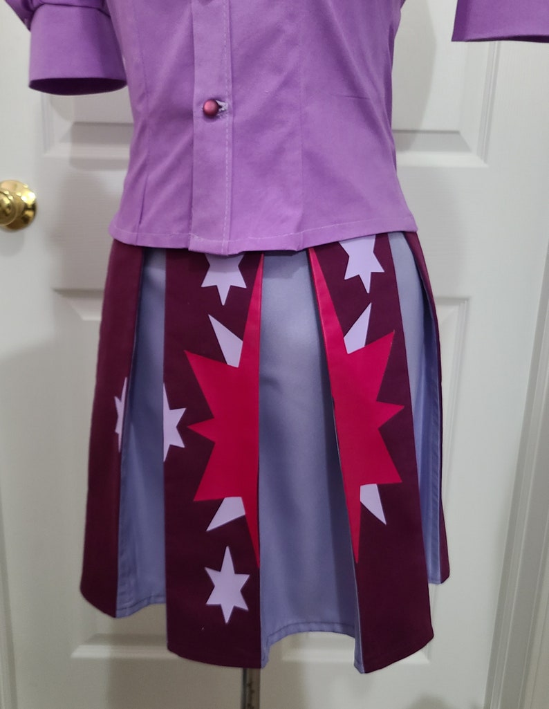 May include: A purple button-up shirt with a pink button and a pleated skirt with a pink and white star pattern. The skirt has a burgundy trim.