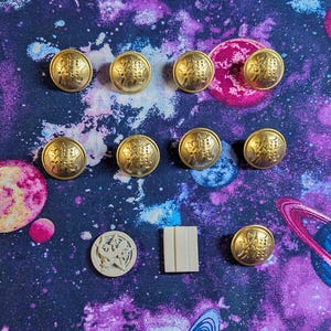 May include: A collection of ten gold-colored buttons with a decorative design. The buttons are arranged in a semi-circle on a purple and blue background with white and pink accents.