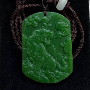 Könnte beinhalten: Grüner Jade-Anhänger mit einem geschnitzten Tiger-Design, an einer braunen Kordel befestigt. Der Anhänger ist rechteckig mit abgerundeten Ecken und wird auf einem schwarzen Samtbeutel präsentiert. Die Kordel ist mit einer weißen Perle befestigt.