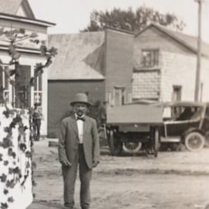 May include: A man in a suit and hat stands in front of a car and a building. He is wearing a bow tie and has a mustache. The car is a vintage model with a wooden body and a canvas top. The building is a two-story structure with a stone facade.