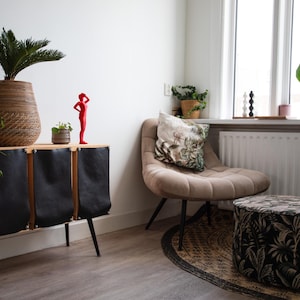 Op de afbeelding: Een moderne woonkamer met een houten kast, zwarte leren deuren en een beige fauteuil. Op de kast staat een grote plant in een gevlochten mand, naast een rood beeldje. De kamer heeft ook een poef met patroon en een groot raam.