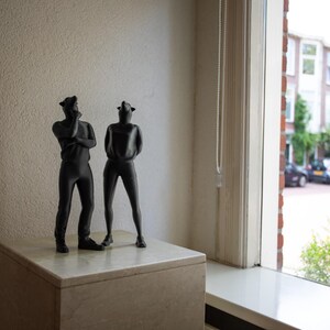 Op de afbeelding: Twee zwarte sculpturen van figuren met dierlijke hoofddeksels staan op een lichtgekleurd stenen blok. Eén figuur heeft de armen gekruist, de andere heeft de handen op de rug. De figuren staan bij een raam.