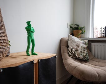 May include: A bright green figurine of a person in a thinking pose stands on a wooden cabinet. The cabinet has black leather panels and silver rivets. A woven basket and a beige chair with a floral pillow are also in the room.