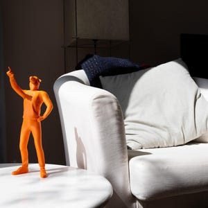 Op de afbeelding: Een oranje beeldje van een persoon die omhoog wijst, staat op een witte tafel. Een witte bank met kussens staat op de achtergrond. De scène wordt verlicht door zonlicht.
