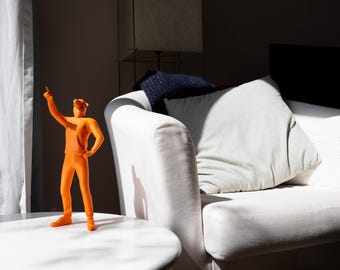 May include: An orange figurine of a person in a pointing pose stands on a white table. The figurine is in front of a white sofa with pillows. The scene is lit by sunlight.