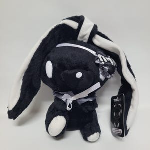 May include: A black and white plush rabbit toy with long ears, white circular eyes, and a white ribbon tied around its neck. The plush features a small white bow on one ear and a black tag attached to its side with a graphic illustration of the rabbit.