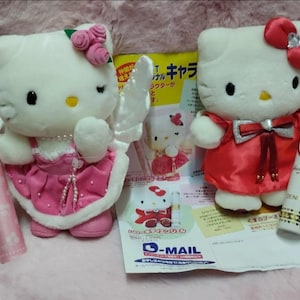 May include: Two Hello Kitty plush toys, one in a pink dress with angel wings and the other in a red dress. A pink cosmetic tube and a white cosmetic tube with gold accents are also present, along with a Japanese magazine.