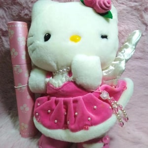 May include: A plush white cat doll with a pink dress, shoes, and a rose hair accessory. The doll has a pearl necklace and a pair of white wings. A pink cylinder with a floral pattern is next to the doll.