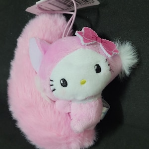 May include: A plush keychain featuring a character with a white face, black eyes, and a yellow nose, wearing a pink hood with a pink bow. The character has a large, fluffy pink tail and a white pom-pom on the side, with a plastic tag attached to the top.