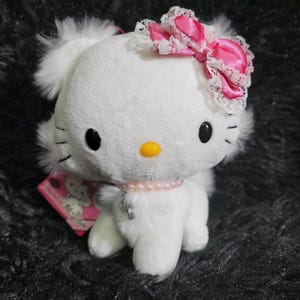 Puede incluir: Un gato de peluche blanco con ojos negros, bigotes y nariz amarilla. Tiene un lazo rosa con ribete de encaje blanco y un collar de perlas rosas con un colgante de llave. Una etiqueta rosa está adjunta.