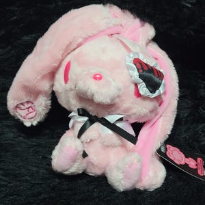 May include: A light pink plush rabbit toy with long ears, featuring a black and white eye patch with a red heart design. The rabbit has a small black and white ribbon bow at its neck and a pink embroidered nose. A black tag is attached to the side.