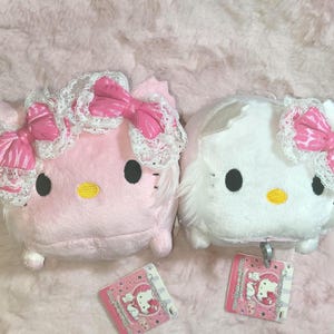 May include: Two plush Hello Kitty dolls, one pink and one white, each with a lace-trimmed bow and a matching pink bow. They have black eyes and yellow noses. Small pink tags are visible below the dolls.