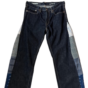 May include: Dark wash denim jeans with patchwork detail on the outer seams. The jeans have a classic five-pocket design and a straight leg fit. The patchwork incorporates various patterned fabrics in shades of blue, white, and green. The tag reads "Gap 1969 Standard 34x34".