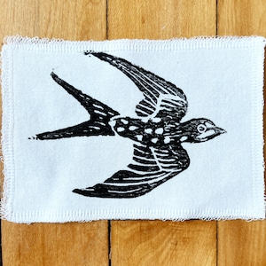 Puede incluir: Una impresión en linóleo en blanco y negro de una golondrina en vuelo, centrada en un parche de tela rectangular blanco. Las alas del ave están extendidas y su cuerpo está detallado con patrones. La tela tiene un borde crudo y deshilachado.