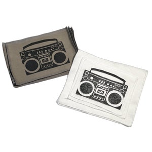 May include: Two square cloth napkins, one gray and one white, each featuring a black graphic of a boombox. The napkins are likely made of cotton or linen and are suitable for kitchen or dining use.