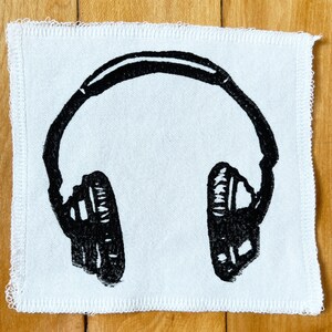 May include: A white square patch with a black graphic of headphones. The headphones are a simple, bold design, with the outline of the headband and earcups clearly visible. The edges of the patch have a stitched border.