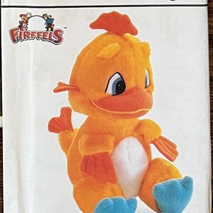 Op de afbeelding: Een zachtoranje Fireffels vogelknuffel met blauwe voeten en een witte buik staat voor een Simplicity knutselpatroon. Het patroon bevat de tekst "crafts/ouvrages" en "Fireffels".
