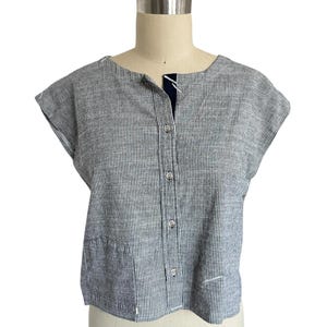 Puede incluir: Una camisa abotonada a rayas grises y blancas con mangas cortas. La camisa tiene un escote redondo y un pequeño bolsillo en la parte inferior delantera. La tela parece ser lino o un material ligero similar.