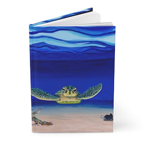 May include: A hardcover journal featuring a vibrant underwater scene. The cover art depicts a sea turtle swimming in deep blue water, with a sandy seabed and a stingray visible. The journal is open, revealing blank white pages.