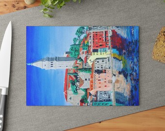 May include: A rectangular cutting board featuring a colorful painting of a coastal town. The artwork depicts buildings in shades of red, yellow, and blue, with a tall white tower. The background is a bright blue sky and water.