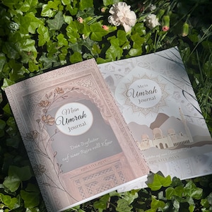 Könnte beinhalten: Zwei Journale mit dem Titel "Mein Umrah Journal" liegen auf einem Bett aus grünen Blättern. Die Journale haben dekorative Einbände mit floralen und architektonischen Designs. Der Text "Dein Begleiter auf einer Reise voll Kheir" ist ebenfalls sichtbar.