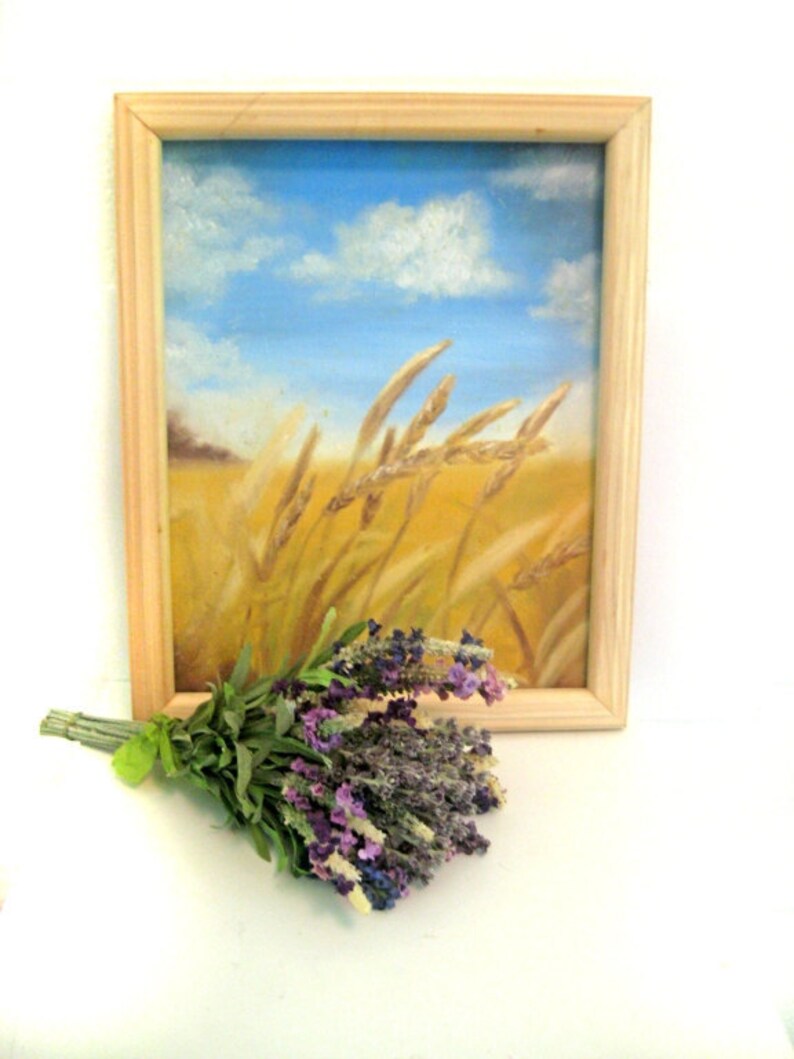 Op de afbeelding: Een ingelijst schilderij van een tarweveld met een blauwe lucht en witte wolken op de achtergrond. Een bosje lavendelbloemen bevindt zich op de voorgrond.