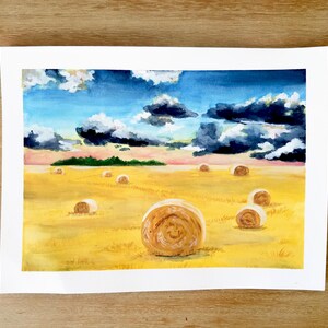 Op de afbeelding: Waterverf schilderij van een veld met hooibalen. De lucht is blauw met witte wolken en wat donkere wolken. Het veld is geel en er liggen verschillende hooibalen verspreid over het veld.