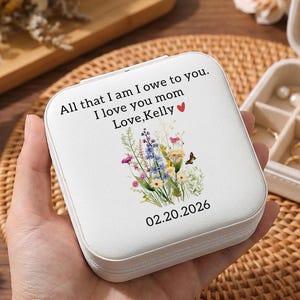 May include: A white, square jewelry box with a floral design and the text "All that I am I owe to you. I love you mom Love, Kelly 02.20.2026". The box is open, revealing compartments with rings, earrings, and necklaces. The box is held in a hand.