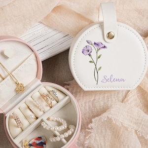 Puede incluir: Un estuche de viaje para joyas rosa y redondo, abierto para mostrar anillos, collares y pulseras. También se muestra un estuche redondo blanco separado con un diseño floral púrpura y el nombre "Selena". Las joyas incluyen detalles dorados y perlados.