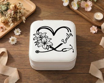 May include: A white square jewelry box with a black heart-shaped design, lilies, and a stethoscope. The name "Emily" is written in script. The box is on a wooden surface with flowers and a candle in the background.