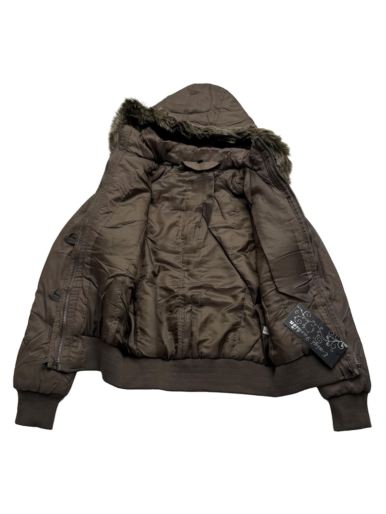 May include: A brown bomber jacket with a hood trimmed with faux fur. The jacket has a zipper closure and ribbed cuffs and hem. The interior lining is a lighter shade of brown. A tag with the text "Ential Fashion" is visible.