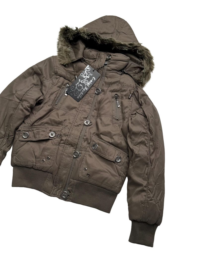 May include: A brown bomber jacket with a fur-lined hood. The jacket features multiple pockets, a zipper closure, and a ribbed hem and cuffs. A tag with the text "E-nigi Fashion" is attached.