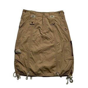 May include: A tan-colored skirt with multiple pockets and adjustable drawstrings at the hem. The skirt features a button and zipper closure, along with decorative straps and stitching details. The skirt has a utilitarian design.