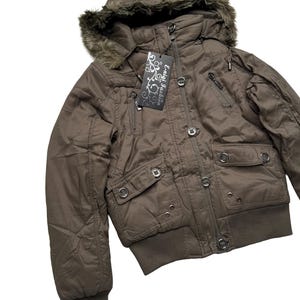 May include: A brown hooded jacket with a faux fur trim around the hood. The jacket has multiple pockets, a zipper closure, and button details. A tag with the text "E-nigi Fashion" is attached. The jacket has a ribbed hem and cuffs.