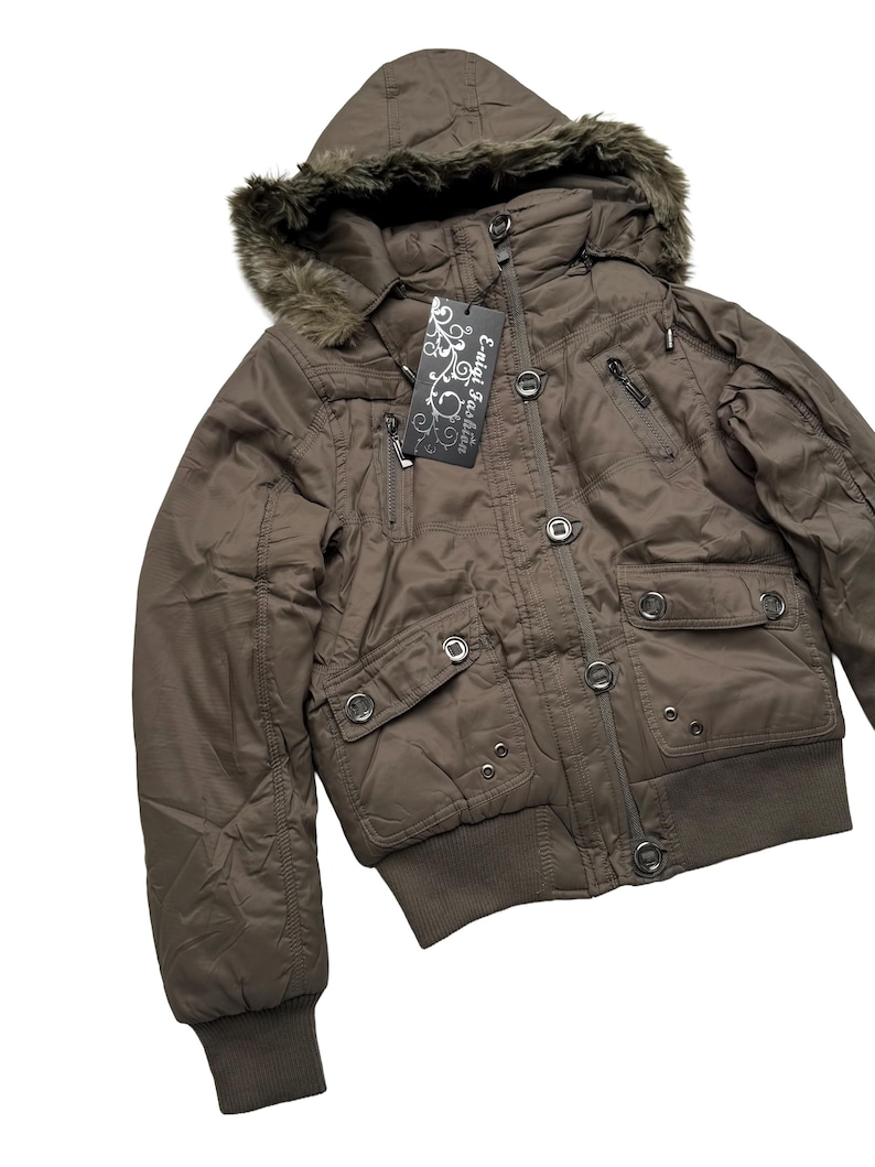 May include: A brown hooded jacket with a faux fur trim around the hood. The jacket has multiple pockets, a zipper closure, and button details. A tag with the text "E-nigi Fashion" is attached. The jacket has a ribbed hem and cuffs.