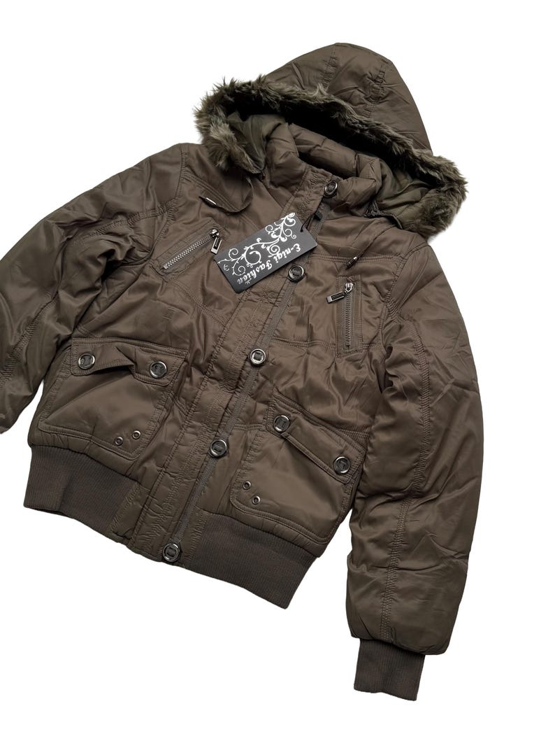 May include: A brown bomber jacket with a fur-lined hood. The jacket has multiple pockets, a ribbed hem and cuffs, and a tag that reads "E-nial Fashion". The jacket is buttoned up and has a quilted design.