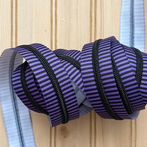 May include: A coiled zipper with alternating purple and black stripes. The zipper teeth are black. A light blue ribbon with vertical stripes is visible on the left side of the image. The background is a light brown wooden surface.