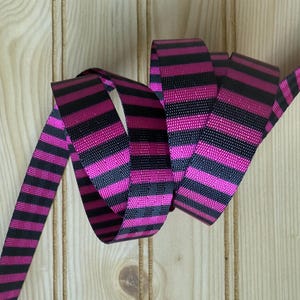 May include: A length of black and hot pink striped ribbon, curled and looped against a light wood grain background. The ribbon is made of a woven material and features evenly spaced stripes. The ribbon is likely used for crafting, gift wrapping, or decorative purposes.