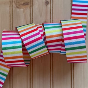 May include: A colorful striped ribbon in a rainbow of hues, including red, pink, orange, yellow, green, and blue, against a light wood background. The ribbon is folded and curved, showcasing its pattern. Ideal for crafting and decorative projects.