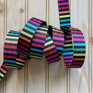 May include: A colorful ribbon with a repeating pattern of horizontal stripes in various colors, including red, orange, yellow, green, blue, and purple, against a black background. The ribbon is curled and lies on a light wood surface.