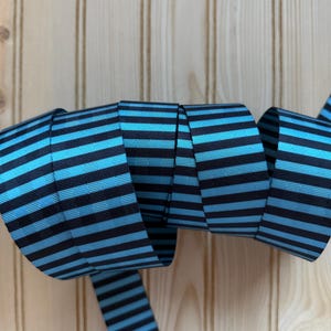 May include: A rolled-up ribbon with alternating horizontal stripes of black and light blue. The ribbon is made of a woven material and is displayed against a light wood background. The ribbon is likely used for crafting or gift wrapping.