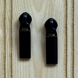 May include: Two black zipper pulls are displayed against a light wood grain background. Each pull has a rounded top with a rectangular body. The pulls are made of a matte black material.