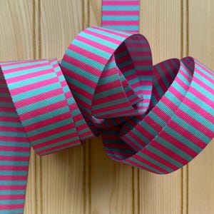 May include: A vibrant ribbon with alternating horizontal stripes of bright pink and turquoise. The ribbon is looped and curled, showcasing its pattern and texture. The background is a light-colored wooden surface.