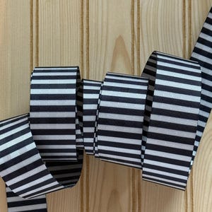 May include: A black and white striped ribbon, curled and displayed against a light wood background. The ribbon's pattern features alternating black and white stripes, creating a bold visual contrast. The ribbon is likely used for crafting or decorative purposes.