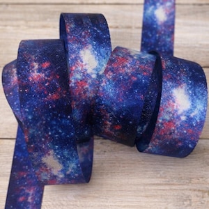 May include: A length of decorative ribbon with a galaxy print. The ribbon features a dark blue background with white, red, and lighter blue accents, creating a cosmic effect. The ribbon is curled and displayed against a wooden surface.