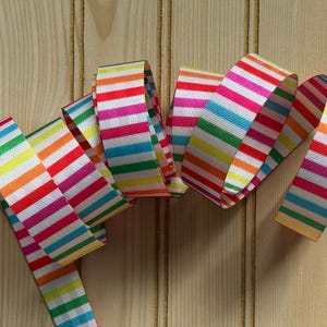 May include: A colorful ribbon with a rainbow striped pattern. The ribbon features horizontal stripes in various colors, including red, pink, orange, yellow, green, and blue. The ribbon is displayed on a light brown wooden surface.