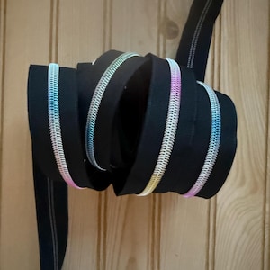 May include: A coiled black zipper with a rainbow-colored metallic tooth. The zipper is shown against a wooden background. The zipper's teeth have a gradient of colors, including blue, pink, and yellow.