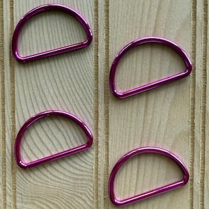 May include: Four magenta-colored D-rings arranged on a light brown wooden surface. Each D-ring is a half-circle shape with a flat bottom. The rings appear to be made of metal and have a shiny finish. The wooden surface has vertical grooves.